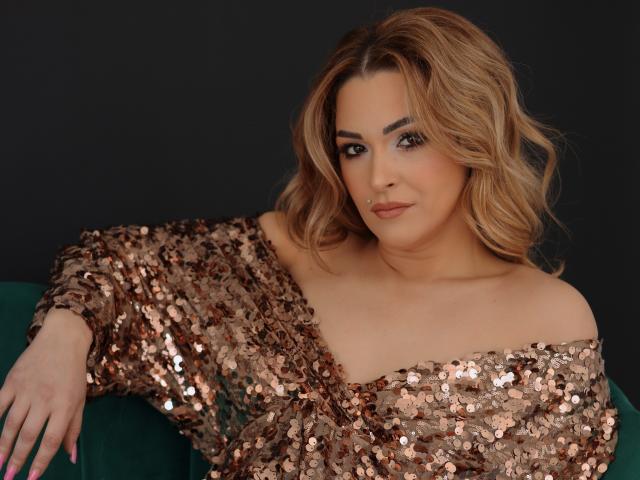 SashaBradley with shoulder-length wavy blonde hair, wearing a sequined off-the-shoulder top, leaning on a dark green couch. Her expression is sultry, with a slight smirk. Shiny sequins, dark background.