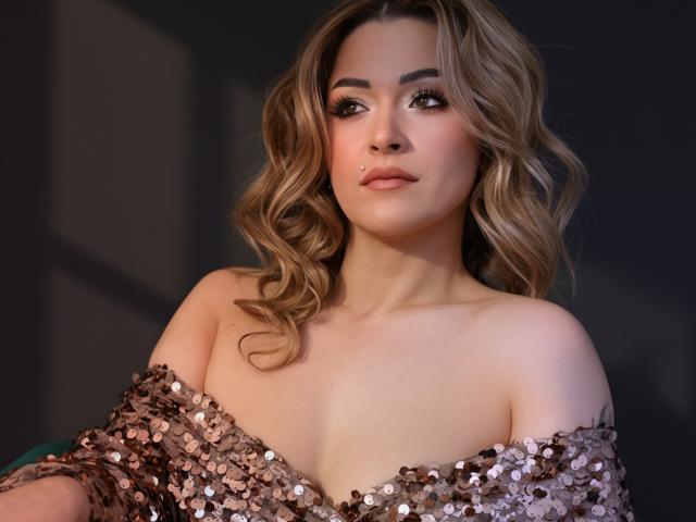 Photograph of SashaBradley, a sexy woman with wavy brown hair, wearing a sparkly off-shoulder sequin dress, showcasing her cleavage. Her expression is confident and slightly seductive, with a dark background.