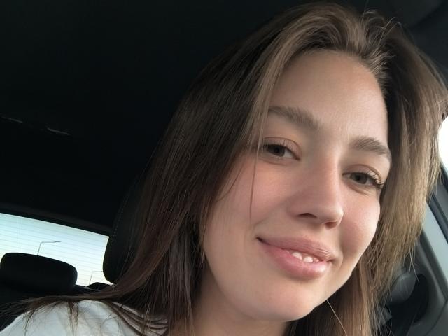 Close-up selfie van ValeriusBertie met een zelfvoldane glimlach, bruine ogen en lichtjes open lippen. Donkerbruin haar, lichte huid, casual witte top. Het licht van het raam werpt een zachte gloed op het gezicht..