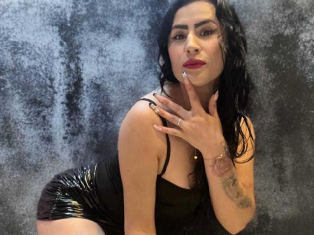 Photographie de DemiMistral, une femme voluptueuse avec de longs cheveux noirs, portant une robe noire en latex moulante, penchée en avant. Sa main sur son cou, ses lèvres rouges, son bras tatoué et son regard intense..