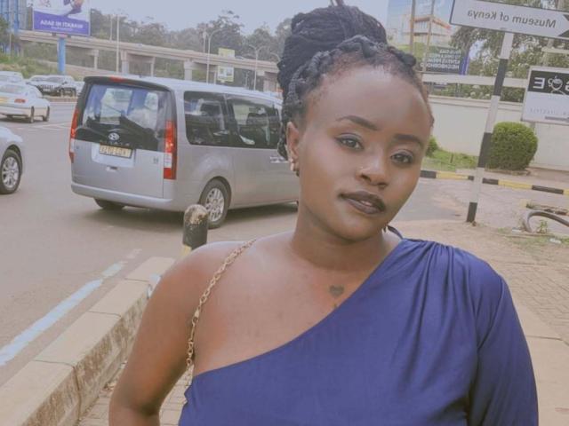 Photographie de BobbieFairy, une femme noire attrayante avec des cheveux tressés, portant une robe bleue à une épaule, debout sur une coin de rue.
une camionnette argentée et une pancarte en arrière-plan. Incroyablement sexy..