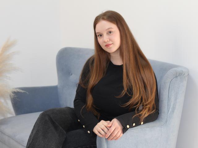ValeriVoom with long, straight brown hair, sitting on a light blue armchair, wearing a black long-sleeve top and black pants, hands resting on chair, subtle sexual tension, relaxed pose.