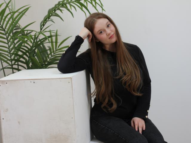 ValeriVoom sits leaning against a white cube, hand in hair, long brown hair, black sweater, dark jeans. Green palm leaves in background. Slight smile, relaxed pose, sexual tension.