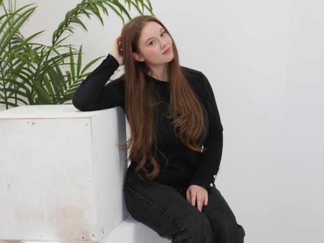 ValeriVoom with long brown hair, sitting against a white wall, wearing a black sweater and dark jeans, hand in hair, leaning slightly, green plants in the background, white planter.