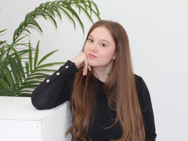 ValeriVoom with long brown hair, black long-sleeve shirt, resting chin on hand, green plant in background, white wall, white and gray borders. Fucking cute, slightly sexy pose.