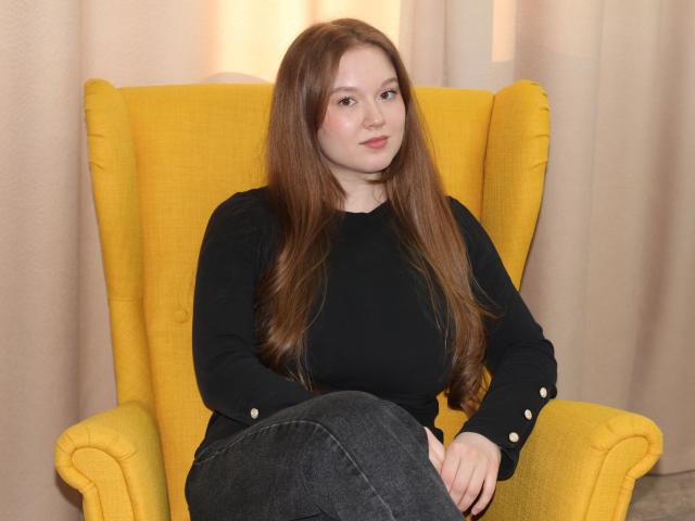 ValeriVoom, a chubby woman with long brown hair, sits in a bright yellow armchair, wearing a black sweater and dark jeans. She's looking directly at the camera with a serious expression.