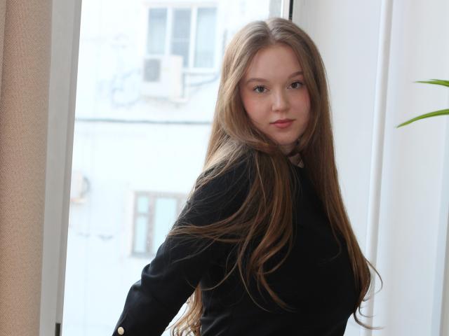 Photograph of ValeriVoom with long brown hair, wearing a black top, standing in front of a bright window. She looks directly at the camera with a slightly smirking expression. Light background.