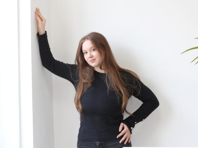 Photograph of ValeriVoom with long brown hair, wearing a tight black long-sleeve shirt, leaning against a white wall with one arm raised, hand on hip, intense gaze, slight smile.