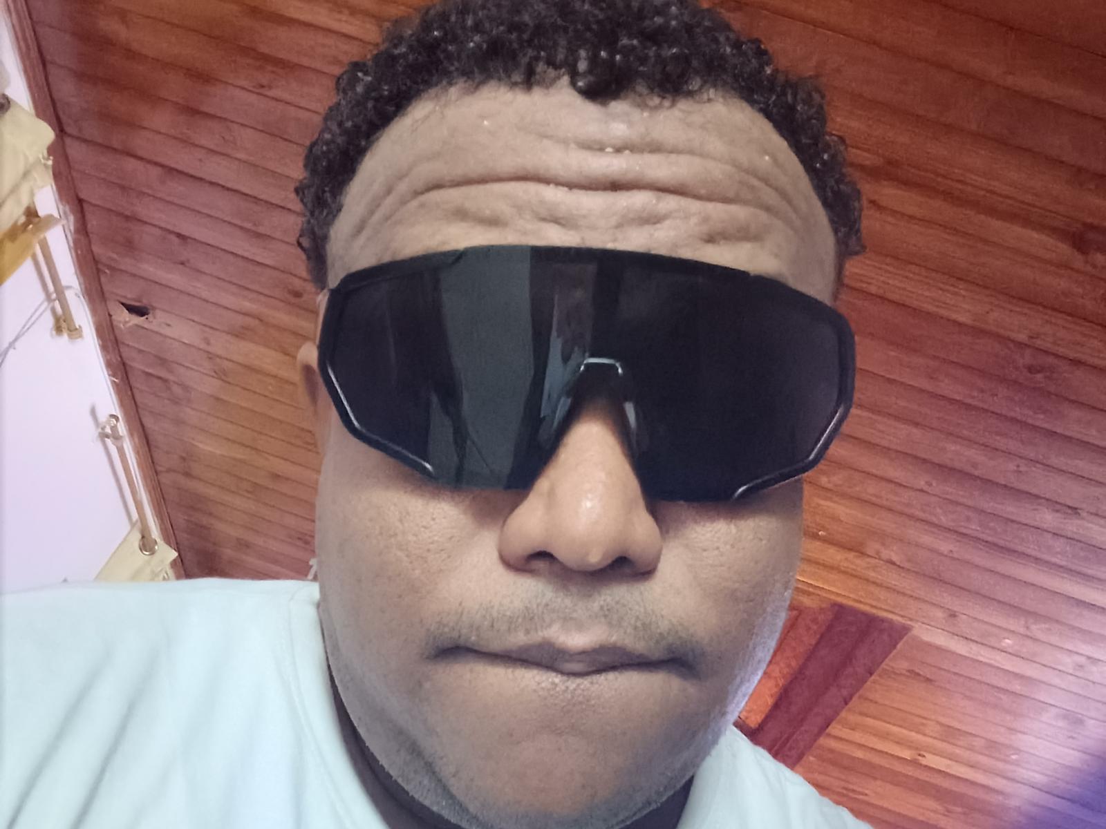 Gros plan de Lpajaro, un homme corpulent avec des cheveux noirs bouclés, portant des lunettes de soleil noires et une chemise blanche. Fond en bois pour le plafond. L'expression sérieuse de Lpajaro, ses yeux ombragés et son front légèrement froncé..