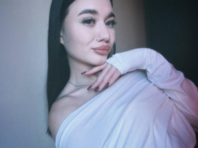 Close-up of AlexaStrei with long black hair, pale skin, and dark eyebrows. She's wearing a white off-shoulder top, hand on chest, smiling slightly, nose ring, dark nails, blue light.