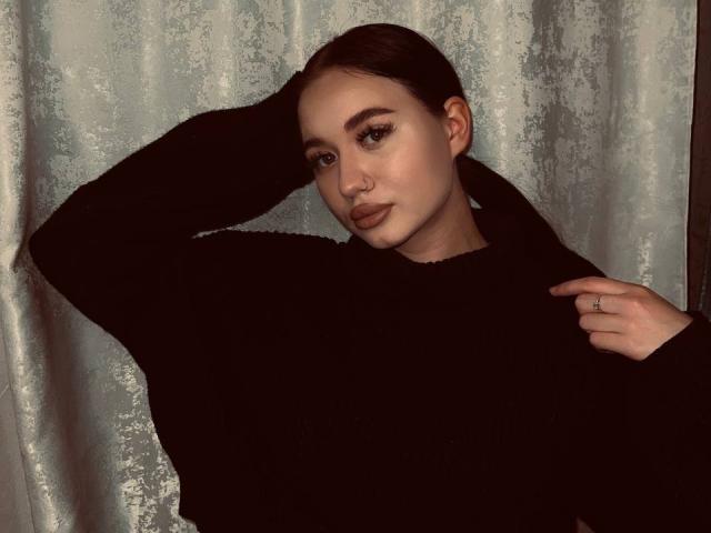 Photograph of AlexaStrei, wearing a black sweater, with one hand behind her head and the other touching her shoulder. She has dark hair, heavy eyeliner, and a nose ring. Silky gray background. Sexual tension.