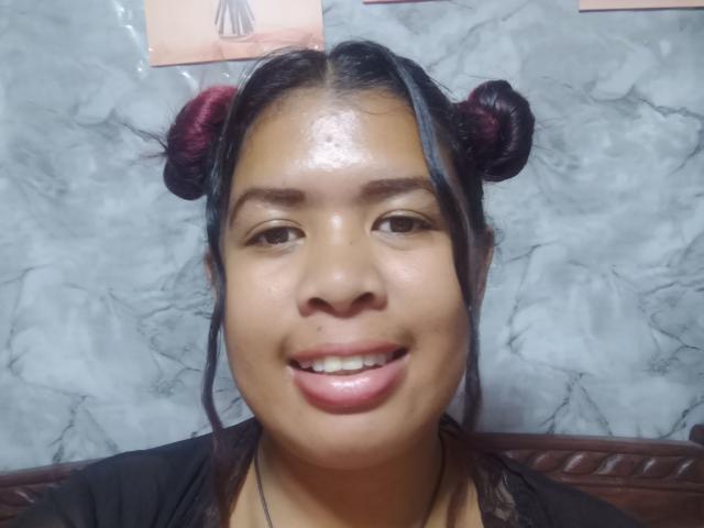 Close-up of MariahSquirting69, an Asian woman with shiny skin, black hair in two buns, red hair ties, smiling with glossed lips. Her face glistens, sitting against a marble wall.