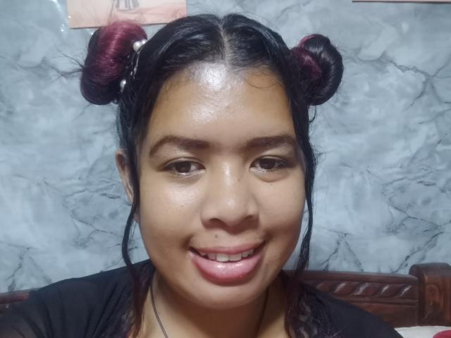 Close-up of MariahSquirting69, an Asian woman with shiny skin, black hair in two maroon-dyed buns, smiling with tongue slightly out. Shiny forehead, marble background, dark top. Sexual tension, slightly provocative.