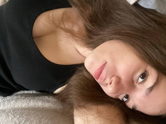 Close-up of KaiaLein with long brown hair, lying on a white surface, wearing a black top, looking up with brown eyes, subtle smile, natural light highlighting her face, sexual tension.