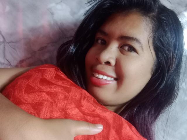 Close-up of MariahSquirting69 with long black hair, smiling with red lipstick, holding a red textured blanket, looking directly at the camera, tan skin, soft light, slight sexual tension.