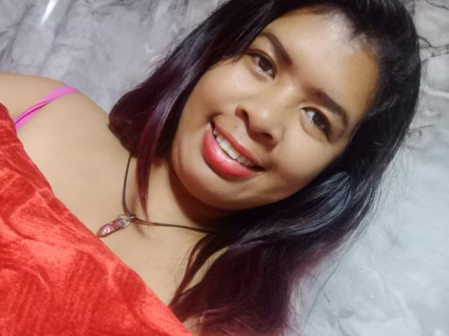 Close-up of MariahSquirting69 with long black hair, smiling with red lipstick. She's wearing a red velvet top, pink bra strap visible. Necklace with pink gem. Bright, direct lighting.