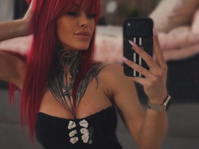 JuicyPeachy – heiß und sinnlich, mit leuchtend rotem Haar, tätowiertem Hals und einem schwarzen, trägerlosen Top mit silbernen Nieten, hält ein Telefon und macht ein Foto. Ein Selfie. Unschärfer Hintergrund eines Schlafzimmers. Armbanduhr am linken Handgelenk. Verdammt sexy..
