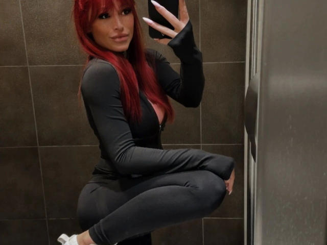 JuicyPeachy: una ragazza dai capelli rossi, formosa, seduta in un bagno con piastrelle scure, che indossa una maglia nera a maniche lunghe e pantaloni aderenti. tenendo uno smartphone, selfie allo specchio, mostrando il décolleté, sexy, sicura di sé, leggero sorriso, calze bianche.