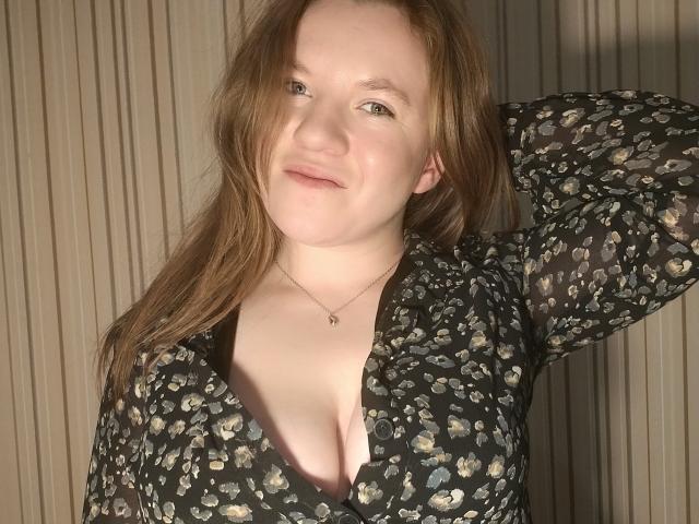 Fotografie von PrettyAhVoron, hellhäutig mit roten Haaren, trägt eine schwarze Bluse mit Blumenmuster, eine Hand ist hinter dem Kopf erhoben. Dekolleté sichtbar, Sonnenlicht betont das Gesicht, Schatten auf der rechten Seite, subtile sexuelle Spannung..