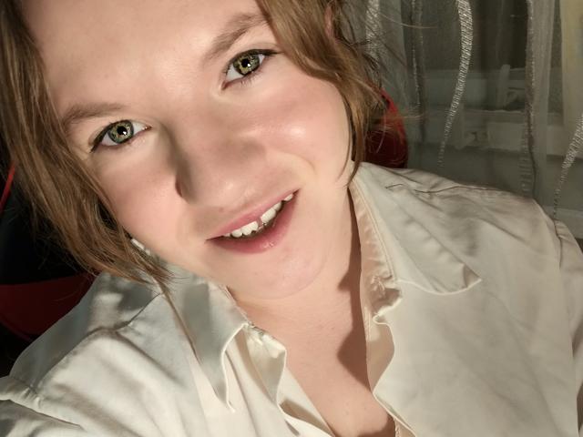 Nahaufnahme eines Fotos von PrettyAhVoron mit heller Haut und braunen Augen, die breit lächelt. Sie trägt ein weißes Hemd mit Knopfleiste, das Sonnenlicht betont ihr Gesicht. Weiche Schatten auf ihren Schultern. Verdammt heiß..