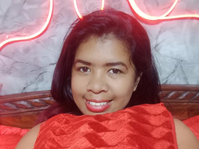 MariahSquirting69 with medium brown skin, black hair, and red lipstick, smiling directly at the camera. She's wearing a red, textured top, with neon lights in the background. Fucking hot.