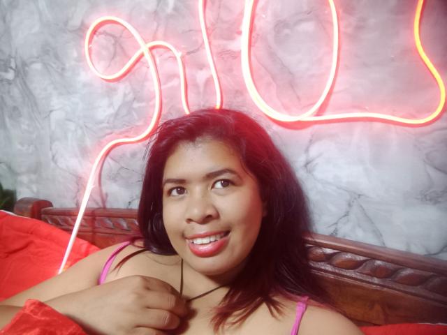 MariahSquirting69, eine dunkelhäutige Frau mit schwarzen Haaren, in einem pinken Tanktop, lächelnd, die ein rotes Tuch hält, unter Neon-Rot-Licht. Kurven hinter ihrem Kopf, Marmorwand, sexy, provokativ, lässig, intime Atmosphäre..