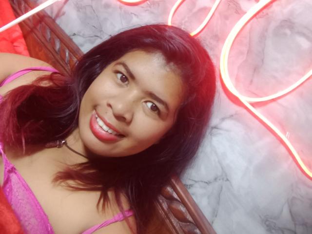 MariahSquirting69 with medium brown skin and black hair, smiling with red lips, wearing a pink lace top, neon red light in the background, leaning forward, visible cleavage, seductive pose.