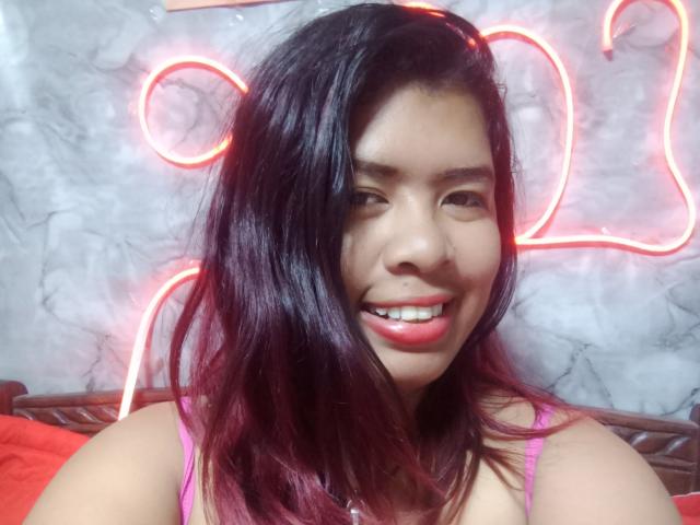 MariahSquirting69 with black hair, smiling, red lipstick, pink top, neon lights in background, red and white hues, sexy expression, casual pose, visible teeth, soft light, intimate atmosphere.