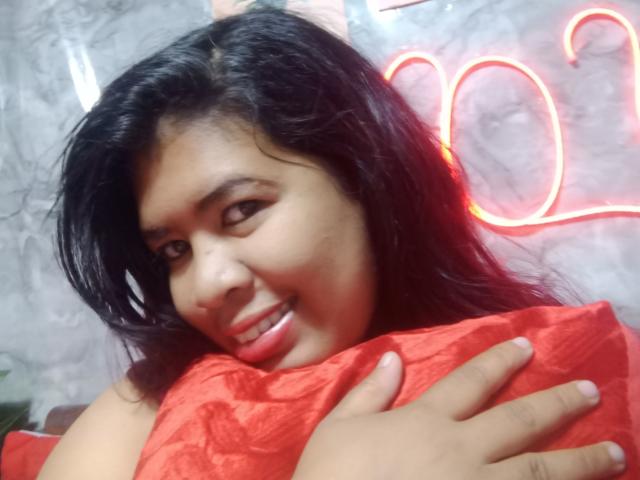 MariahSquirting69 with black hair, holding a red pillow, smiling suggestively. Neon heart outline in background, red lighting on face. Hands gripping pillow, slight shadow on cheek. Casual, sexy vibe.