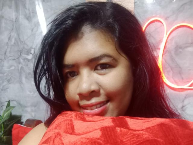 Close-up of MariahSquirting69, an Asian woman with long black hair, smiling with red lips, holding a bright red fabric against her chest, neon lights in the background. Sexual tension, seductive gaze.