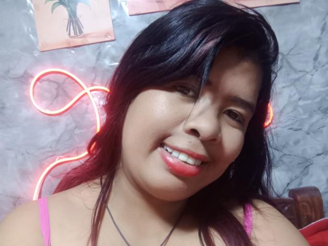 Close-up of MariahSquirting69 with dark straight hair, smiling with red lipstick, wearing a pink top. Neon lights in the background, including a red loop and white light. Casual, sexy vibe.