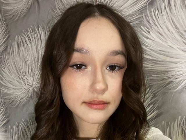 Close-up de AnitraAmphy, uma jovem com pele clara, cabelo ondulado escuro e batom vermelho, olhando diretamente para a câmera. A expressão dela é calma, com um toque de tensão sexual. O fundo apresenta texturas prateadas e fofas..