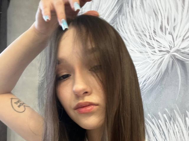 Close-up foto van AnitraAmphy, een jonge vrouw met lang bruin haar, licht open lippen en een kleine glimlach. Ze streelt haar haar en laat een tatoeage op haar linkerarm zien. Witte, draaiende achtergrond..