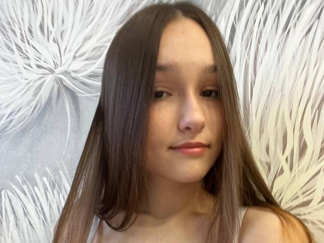 Fotografía de AnitraAmphy con cabello largo y lacio de color marrón, piel clara y una sonrisa sutil. Ella lleva una camiseta blanca sin mangas y está de pie frente a un fondo blanco con textura y un estampado floral. Increíblemente atractivo..