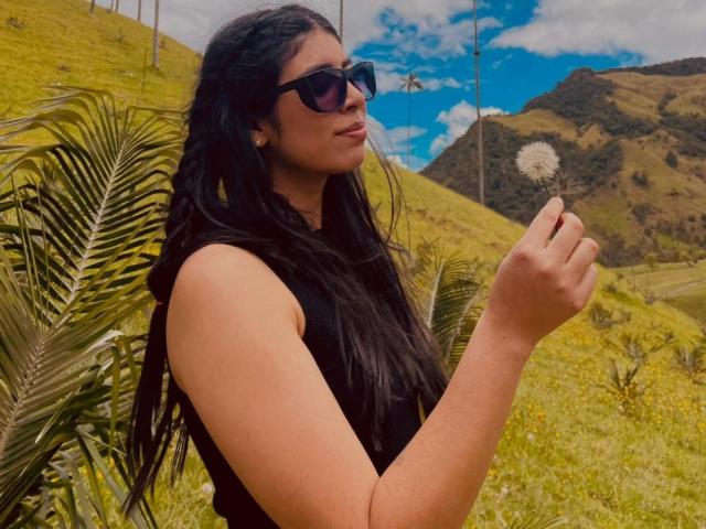 MiluSky, con cabello largo y negro, usando gafas de sol oscuras, sosteniendo una semilla de diente de león, de pie en un paisaje soleado y montañoso. con hojas de palma. El fondo muestra hierba amarilla y cielo azul. Increíblemente atractivo..