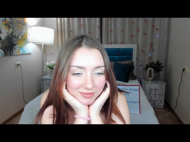 Fotografia ravvicinata di AllaMiller, una donna dalla pelle chiara con lunghi capelli castani, che sorride direttamente verso la fotocamera. Lei appoggia il mento sulla mano, con una fonte di luce intensa sul viso. Sfondo di una camera da letto con tende, lampada e letto. Tensione sessuale, attraente, incredibilmente sexy..