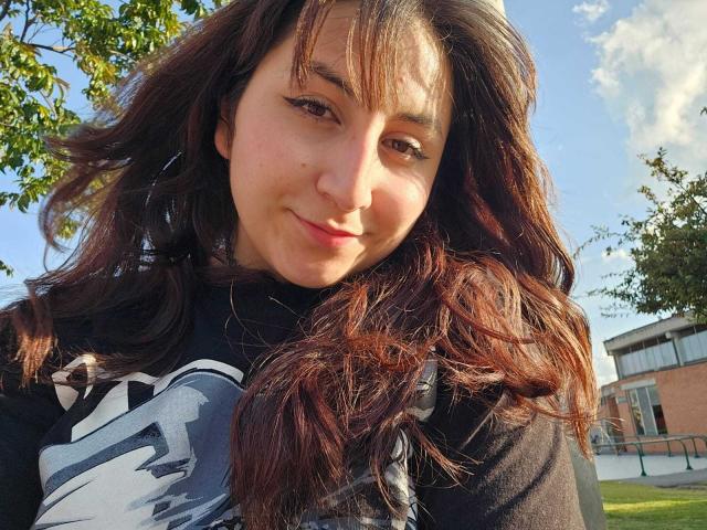 Nahaufnahme von MaddieLesYoux, einer Frau mit langen, braunen Haaren und Pony, die leicht lächelt und ein schwarz-weiß gemustertes Kleid trägt. oben, Sonnenlicht beleuchtet ihr Gesicht, Baum und Gebäude im Hintergrund..