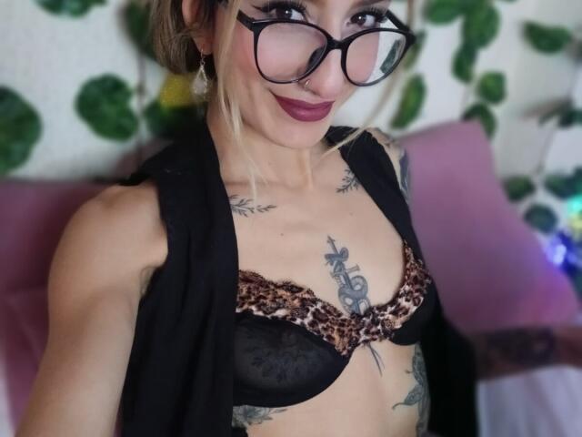 Fotografia ravvicinata di Kaiix, una donna esile con tatuaggi, che indossa un reggiseno nero in pizzo e con stampa leopardo, una giacca nera aperta e occhiali. Ha un rossetto scuro e un sorriso sicuro. Lo sfondo è sfocato..