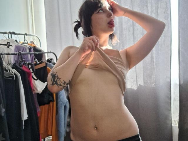 KenyattaConsigli, a pale-skinned woman with dark hair, lifts her beige tank top, exposing small breasts and a tattoo on her left arm. She's looking up, mouth slightly open. Sunlight filters through curtains.
