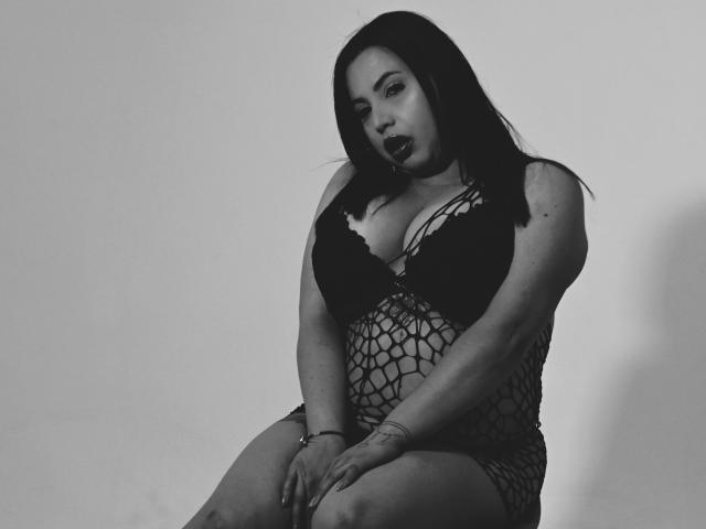 Fotografia em preto e branco de Nahomi Rosse sentada com as pernas cruzadas, vestindo um macacão de meia-calça que revela grandes áreas do corpo. seios e um decote. Ela olha de forma sedutora, os lábios levemente entreabertos, o cabelo escuro caindo..