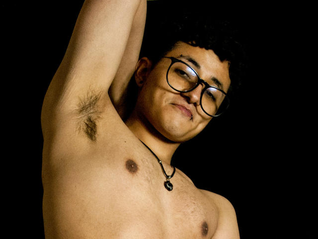 Photographie d'Adoni Baskerville, torse nu avec des poils sous les aisselles, levant le bras, portant des lunettes, un collier noir, avec des cheveux foncés. Peau claire, fond noir.
cock apparent, tension sexuelle, regard intense, pose décontractée.