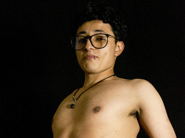 Adoni Baskerville, torse nu, avec des cheveux noirs bouclés, portant des lunettes à monture noire et un collier avec un petit pendentif, a une expression confiante et un mamelon visible, sur un fond sombre. Extrêmement séduisant..