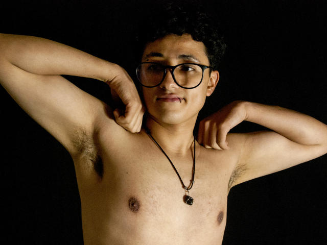 Adoni Baskerville, sin camisa y con los brazos levantados, mostrando las axilas, lleva gafas y un collar. Fondo oscuro, tono de piel medio, pezones pequeños, ligera sonrisa. Imagen fotográfica con iluminación de alto contraste..