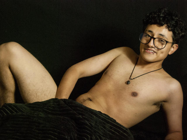 AdoniBaskerville, un homme asiatique torse nu avec des lunettes, allongé sur le dos, les jambes écartées, portant un pantalon noir à rayures, une chaîne autour du cou, fond sombre, sourire narquois, éclairage mettant en valeur sa poitrine lisse et son pénis. Pose décontractée et séduisante.