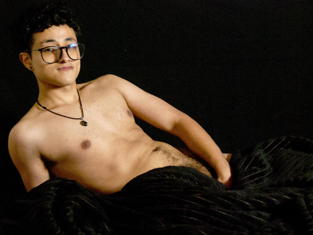 Adoni Baskerville, ohne Hemd, mit schwarzen Haaren und Brille, lehnt sich gegen einen schwarzen Hintergrund. Er trägt eine Halskette und hat eine entspannte Pose, wobei sein Penis sichtbar ist und die schwarzen Hosen teilweise heruntergezogen sind..