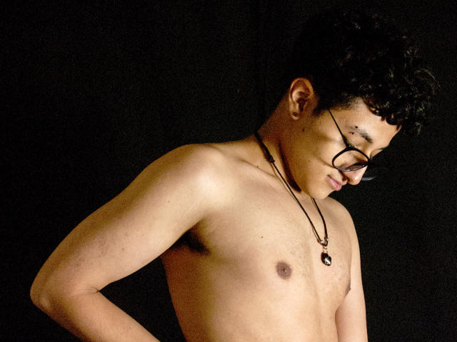 Adoni Baskerville, zonder shirt, met kort krullend zwart haar, een bril en een ketting, kijkt naar beneden met een lichte glimlach, zwarte achtergrond, zacht licht dat het bovenlichaam accentueert, lichte schaduw op het gezicht..