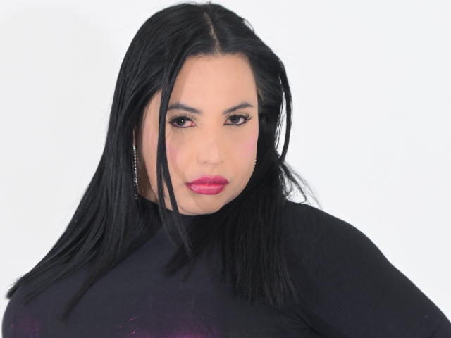 Nahaufnahme eines Fotos von NahomiRosse, einer Frau mit langen, schwarzen Haaren, die ein schwarzes Oberteil, roten Lippenstift und Hängeohrringe trägt. Sie hat einen verführerischen Blick, einen intensiven Ausdruck und leicht geöffnete Lippen..