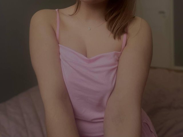 Photograph of Sambucka in a tight, pink ribbed dress with thin straps, showing cleavage. Sambucka's arms are crossed over her chest, brown hair partially visible. Soft lighting, slight shadow.