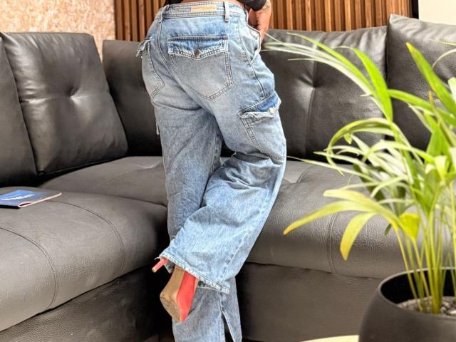 Close-up of NenaBlack's lower body, wearing frayed blue jeans with a red patch on the left ankle, leaning over a black leather couch. Bright green plant in the foreground.