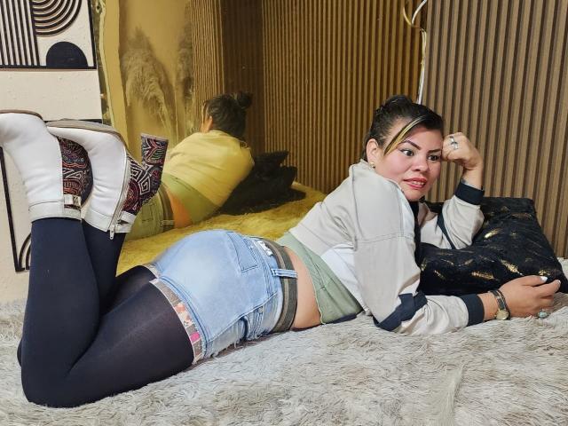 KiaraXMoon è sdraiata a pancia in giù su un tappeto bianco e soffice, indossando una giacca bianca, pantaloncini di jeans e calze nere che arrivano sopra la coscia. con le gambe sollevate e i piedi infilati in calze bianche. Il suo pene è visibile attraverso i suoi pantaloncini..